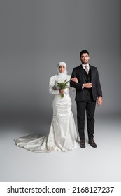 full length of groom in suit standing with muslim bride with wedding bouquet of calla lily flowers on grey