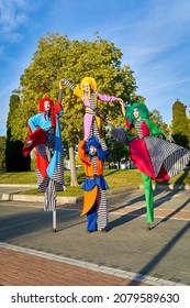 Full length group of happy clowns with painted faces in colorful costumes holding hands and having fun while standing on stilts in park, on sunny day