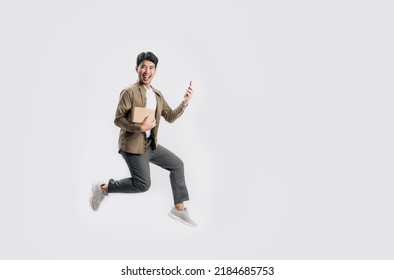 Full Length Handsome Asian Man Holding Box And Use Smartphone He Is Jumping In Air On White Background. Young Male Happy Excited And Using Mobile Phone With Shopping Online Concept.