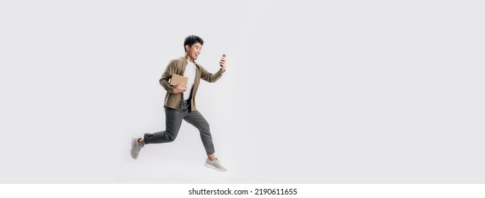 Full Length Handsome Asian Man Holding Box And Use Smartphone He Is Run And Jumping In Air On Banner White Background. Young Male Happy Excited And Using Mobile Phone With Shopping Online Concept.