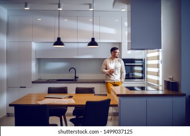 Full length of handsome Caucasian man dressed casual leaning on kitchen counter, drinking coffee and looking trough window. Modern kitchen interior. - Powered by Shutterstock