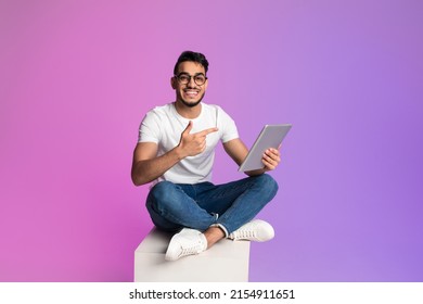 Full length handsome millennial Arab man pointing at digital tablet  checking new app in neon light  copy space  Young middle Eastern guy sitting cross legged and touch pad  working online