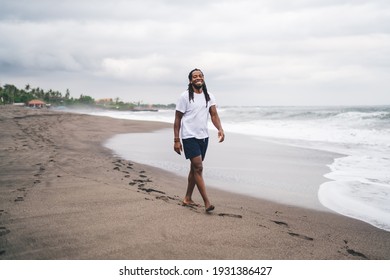 Full length happy African American male in casual wear smiling and looking at camera while walking on sandy beach near waving sea - Powered by Shutterstock