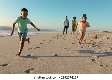 Full length of happy african american children running against parents walking at beach on sunny day. unaltered, family, lifestyle, togetherness, enjoyment and holiday concept.