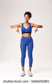 Full Length Of Happy African-american Fitness Coach In Blue Sportswear, Pointing Fingers Down And Smiling, Recommend Workout Gear, Showing Logo Or Sports Promo, White Background