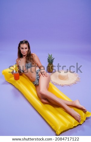Full length of happy beautiful caucasian woman who wears in bikini lyes on inflatable yellow matress while holds cocktail on purple background . Summer concept. Travel concept. Relax concept 