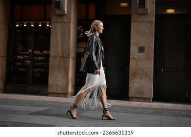 Full length happy blonde woman walking city street in stylish clothes. Young female model smiling, wears white fringed skirt, black leather jacket and sandals. - Powered by Shutterstock