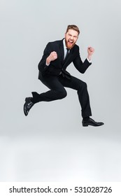 Full Length Happy Business Man In Black Suit Jumping In Studio And Looking At Camera With Open Mouth. Isolated Gray Background