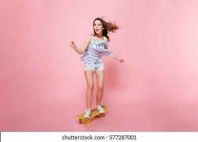 Full Length Of Happy Carefree Young Woman Riding On Skateboard Over Pink Background