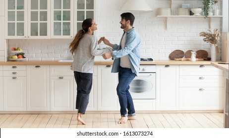 Full Length Happy Family Couple Dancing Barefoot On Wooden Heated Floor In Modern Light Kitchen. Overjoyed Mixed Race Young Married Spouse Enjoying Leisure Weekend Time Activity Together At Home.