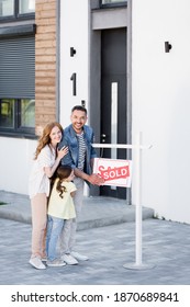 Full Length Of Happy Family Hugging While Looking At Camera Near Sign With Sold Lettering And House