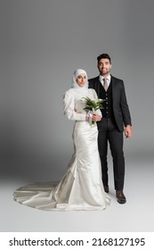 full length of happy groom in suit near muslim bride with wedding bouquet on grey