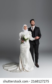 full length of happy groom in suit holding hand of muslim bride with wedding bouquet of calla lily flowers on grey