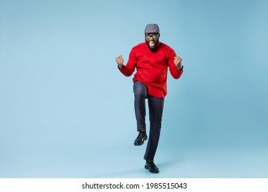Full length happy joyful young bearded african american man in casual red shirt cap eyeglasses standing doing winner gesture clenching fists isolated on pastel blue color background studio portrait