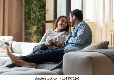 Full length happy loving young couple lying on huge comfortable sofa, holding cups of hot beverages. Smiling family spouse relaxing together on cozy couch with tea coffee, enjoying romantic time.