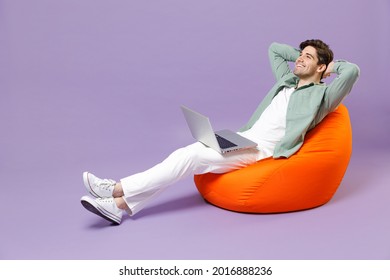 Full Length Happy Man 20s In Casual Mint Shirt White T-shirt Sitting In Orange Bean Bag Chair Hold Hands Folded Behind Neck Use Laptop Pc Computer Isolated On Purple Color Background Studio Portrait