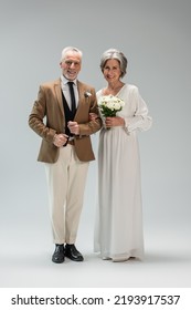 Full Length Of Happy Mature Bride In Wedding Dress Holding Bouquet And Standing With Middle Aged Groom In Suit On Grey