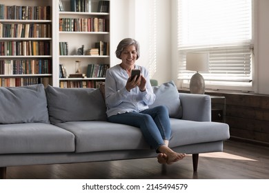 Full length happy middle aged 50s woman using smartphone apps, sitting on comfortable sofa at home, enjoying communicating in social network, playing games. shopping or holding video call conversation - Powered by Shutterstock