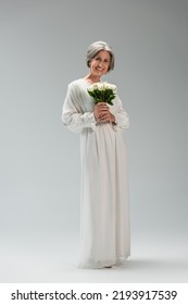 Full Length Of Happy Middle Aged Bride In White Dress Holding Wedding Bouquet On Grey