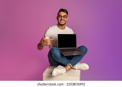 Full length happy millennial Arab man sitting cross legged and laptop computer  pointing at blank screen  showing free space for mockup in neon light  Online advertisement concept