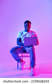 Full length of happy millennial black guy with laptop pc sitting on chair, studying or working online in neon light, copy space. Cheery African American man having remote business meeting on web - Shutterstock ID 2198568243