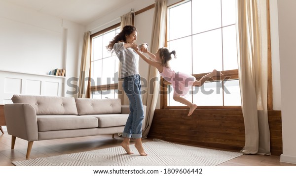 Full length happy mother holds hands spinning\
carefree adorable little daughter listen music moving in modern\
interior light cozy living room. Active funny games, lively play\
have fun together at home