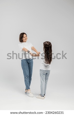 full length of happy mother with tattoo on hand and short hair holding hands with preteen daughter while standing together in white t-shirts and blue denim jeans on grey background
