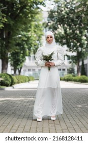 full length of happy muslim bride with diamond ring on finger holding wedding bouquet