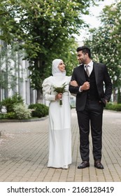 full length of happy muslim bride in wedding hijab and white dress holding bouquet near groom outside
