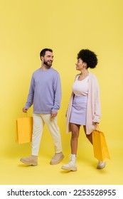 full length of happy and stylish interracial couple walking with shopping bags and looking at each other on yellow background - Shutterstock ID 2253528955