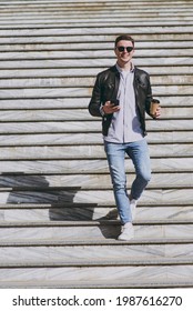 Full Length Happy Stylish Young Man In Leather Jacket Glasses Walk In City Downtown Go Down Concrete Steps Hold Mobile Cell Phone Takeaway Craft Paper Cup Drink Coffee To Go Urban Lifestyle Concept