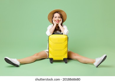 Full length happy traveler tourist woman in casual clothes hat sitting with outstretched legs near suitcase isolated on green background Passenger travel abroad weekends Air flight journey concept.