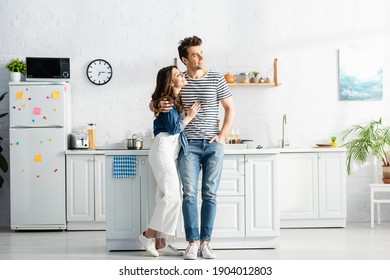 full length of happy woman hugging cheerful man standing with hand in pocket in modern kitchen