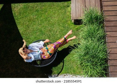 Full Length Of Happy Woman Wearing Straw Hat And Beachwear While Relaxing On A Lounge Chair Outside. Attractive Woman Enjoying Summer Weather. Shot From Above. 