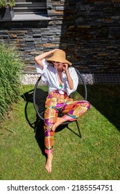 Full Length Of Happy Woman Wearing Straw Hat And Beachwear While Relaxing On A Lounge Chair Outside. Attractive Woman Enjoying Summer Weather. Shot From Above. 