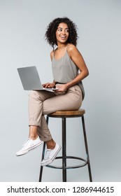 Full length of a happy young african woman casually dressed sitting on a chair isolated over gray background, working on laptop computer