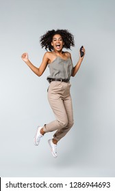 Full Length Of A Happy Young African Woman Casually Dressed Jumping Isolated Over Gray Background, Holding Mobile Phone