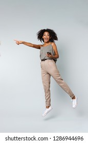 Full length of a happy young african woman casually dressed jumping isolated over gray background, pointing at copy space