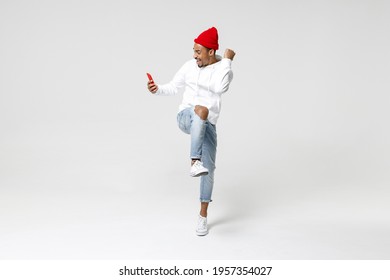 Full length of happy young african american man 20s in casual streetwear hoodie standing using mobile cell phone typing sms message doing winner gesture isolated on white background, studio portrait