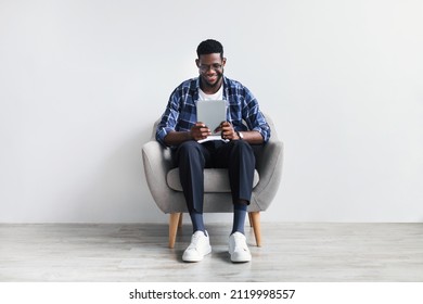 Full length of Happy young black man with tablet sitting in armchair, working or studying online, having video chat against white studio wall. African American guy communicating on web via touch pad