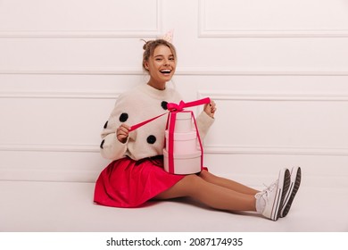 Full Length Happy Young Caucasian Woman Sitting On Floor Unpacking Gift Boxes Indoors. Blonde With Her Hair Pulled Back, Wearing Sweater And Skirt. Birthday Concept