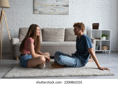 Full Length Of Happy Young Couple Sitting On Carpet And Looking At Each Other In Living Room