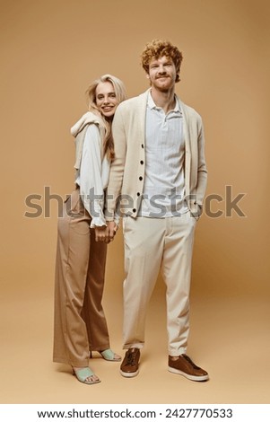 full length of happy young couple in trendy casual clothes holding hands looking at camera on beige