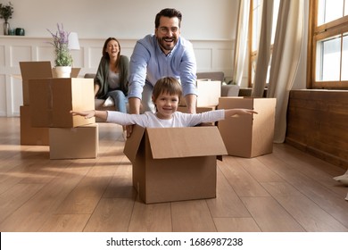Full length happy young father in glasses pushing carton box with small kid son in new living room. Overjoyed family couple having fun with little child, celebrating moving into flat apartment.
