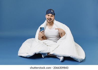 Full Length Happy Young Fun Man In Pajamas Jam Sleep Mask Rest Relax At Home Sit Wrap Under Cover Blanket Duvet Point Finger On Mobile Cell Phone Isolated On Dark Blue Background Night Bedtime Concept