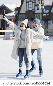 full length of happy young man in winter hat behind smiling girlfriend with outstretched hands on ice rink