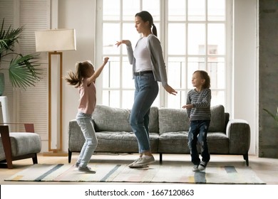 Full length happy young mother dancing with small children siblings in living room. Small energetic kids brother sister having fun with positive nanny together at home, enjoying daycare time.