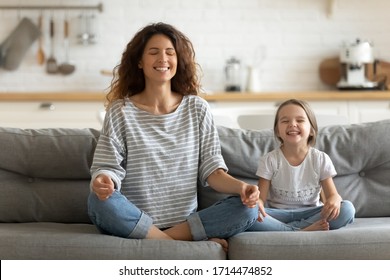 Full Length Happy Young Nanny Mom Sitting On Sofa In Lotus Pose, Teaching Small Daughter Yoga Breathing Exercise. Friendly Sincere Two Female Generations Family Relaxing On Couch, Healthcare Concept.