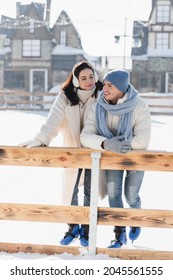 full length of happy young woman in ear muffs looking at cheerful man in winter hat leaning on wooden border on ice rink