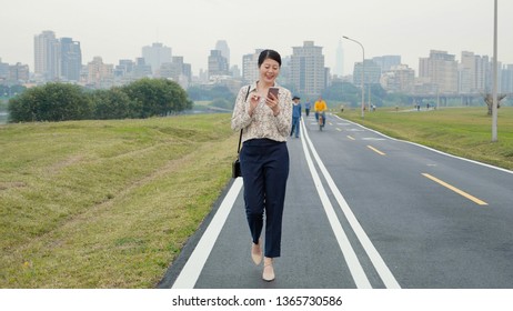 full length hard working woman designer walking on road in city urban park on weekends after work. people workout ride bike with skyscraper in background. lady worker using mobile phone smiling happy